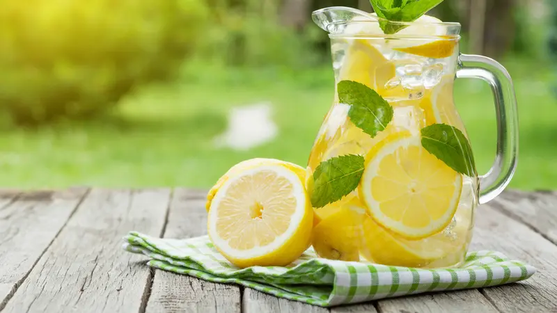 Segelas air lemon segar berada di atas meja dengan latar belakang taman hijau, menampilkan irisan lemon dan dedaunan mint yang menambahkan sentuhan segar